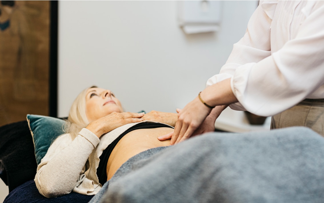 A women likes on the bed with her stomach showing. A practitioner lays their hands on her stomach